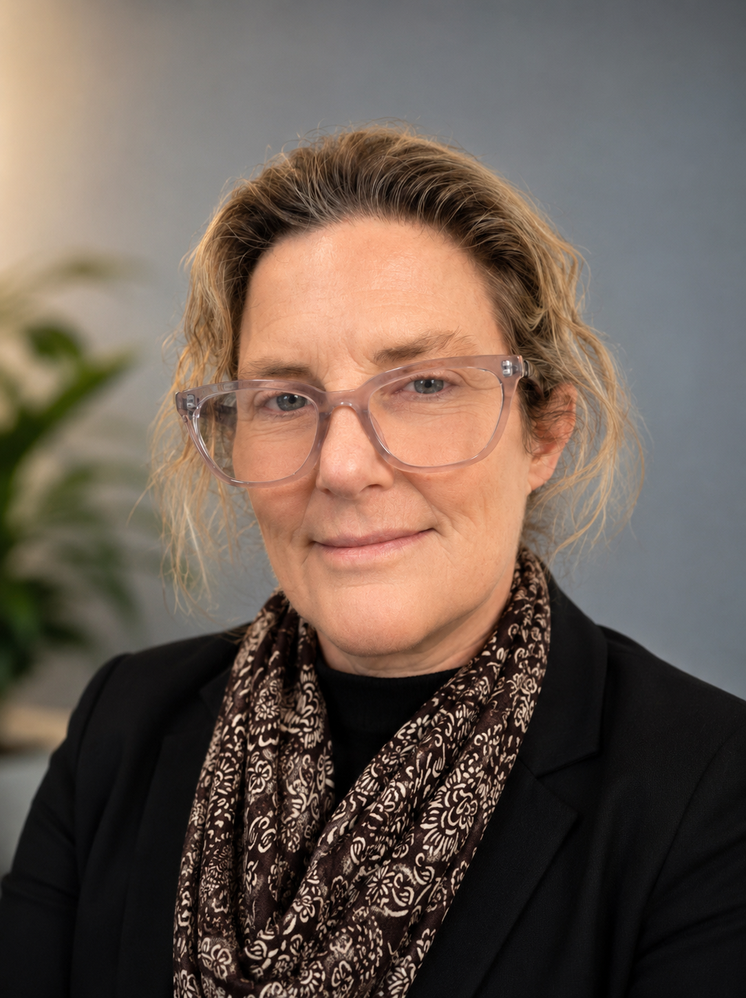 Professional head-and-shoulders photo of a woman with light pink glasses, wearing a black blazer and patterned scarf against a neutral blue-gray backdrop