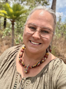 Smiling woman with glasses wearing a brown beaded necklace outdoors on a sunny day.