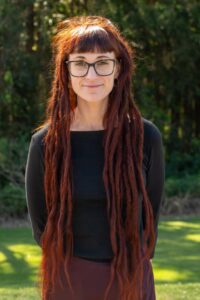 Portrait of a woman with long red dreadlocks and black-rimmed glasses outdoors in a grassy park.