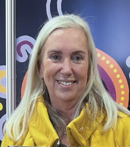 Smiling blonde woman wearing a yellow scarf, with a bunny ears headband, in front of a colorful backdrop.